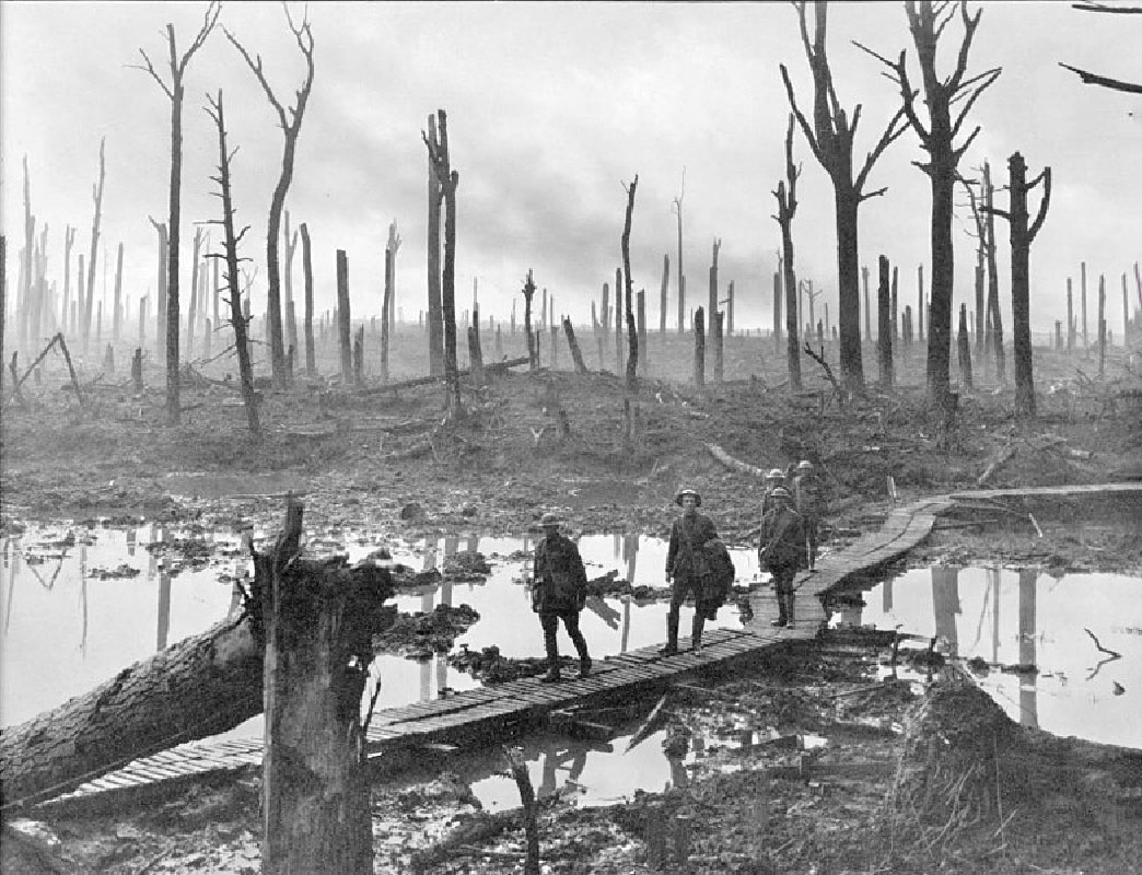 In the background of the photo, several trees are damaged, and mostly burned. The earth is bare. In the foreground, soldiers walk across a raised walkway that cuts through mudholes and fallen trees.