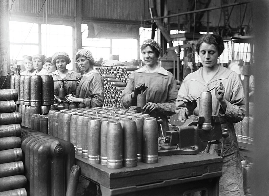 Women stand in a room full of stacked artillery shells. The women stand on one side of table and assemble the shells.