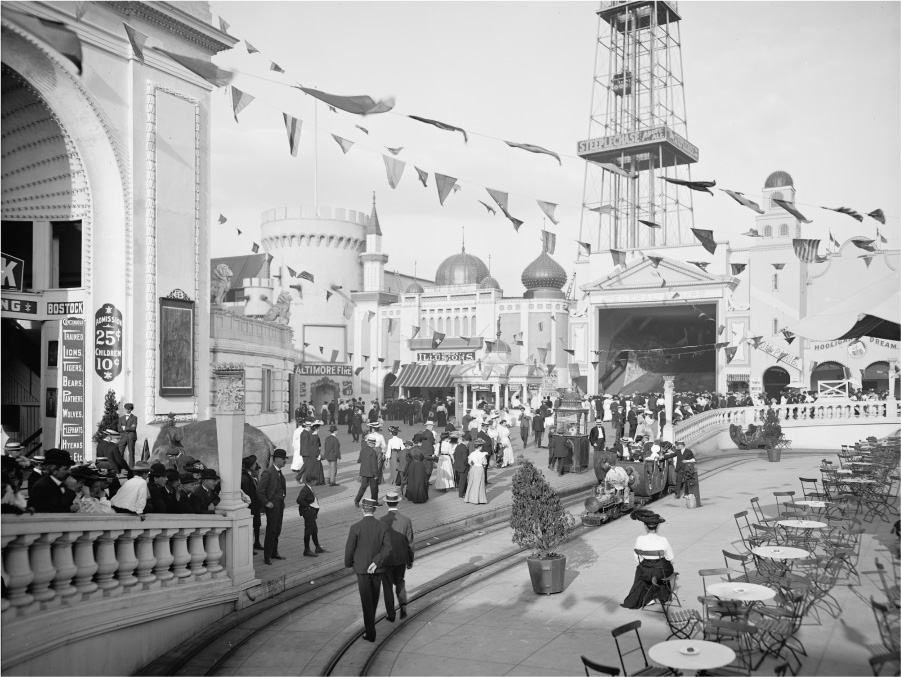 This photograph shows a crowd of people visiting the Dreamland amusement park. All the men are wearing suits and hats. The women are wearing long blouses, skirts, and hats. The children are dressed like adults. Signs list attractions and prices. Several people are riding a miniature train.