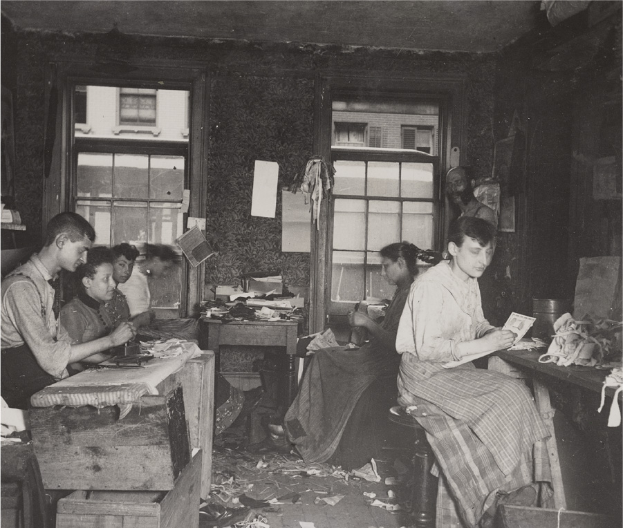 This photograph shows men and women working together in a small room. The room is cluttered, and trash covers the floor. Other buildings are visible through windows in the background.