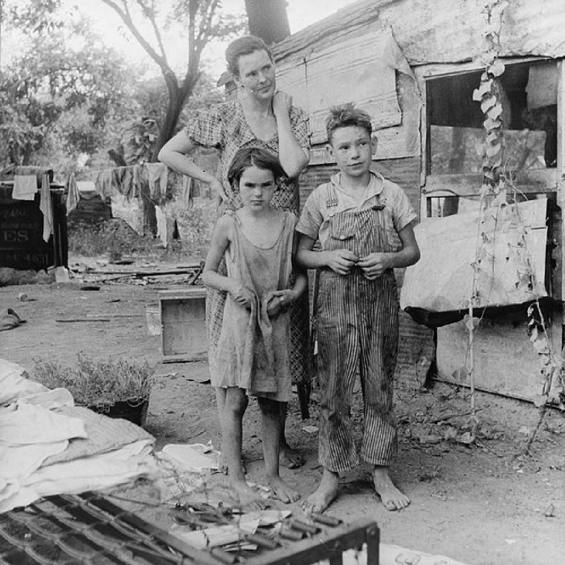 The photograph captures a woman and two children. They are barefoot and their clothes look dirty. Trash is visible nearby.