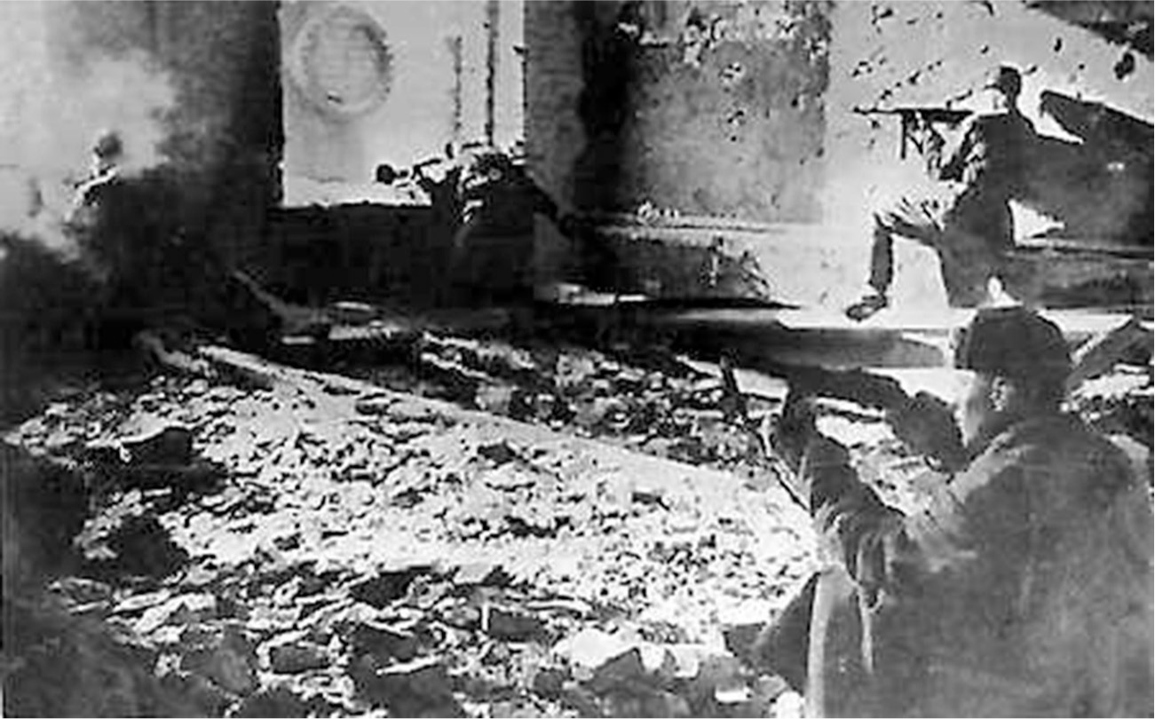 This is a black and white picture of a war scene. There is a scarred wall in the background and a wall with two windows on the left. The middle and bottom left of the picture show rubble all over the ground. There are four soldiers, one positioned at the top left, squatting and looking out a window, with smoke around him, one kneeling at the top middle by a window, one at the top right on one knee along a wall, and one at the bottom right of the picture sitting on the floor. They all have military uniforms on with helmets and are facing left, aiming their rifles toward the left.
