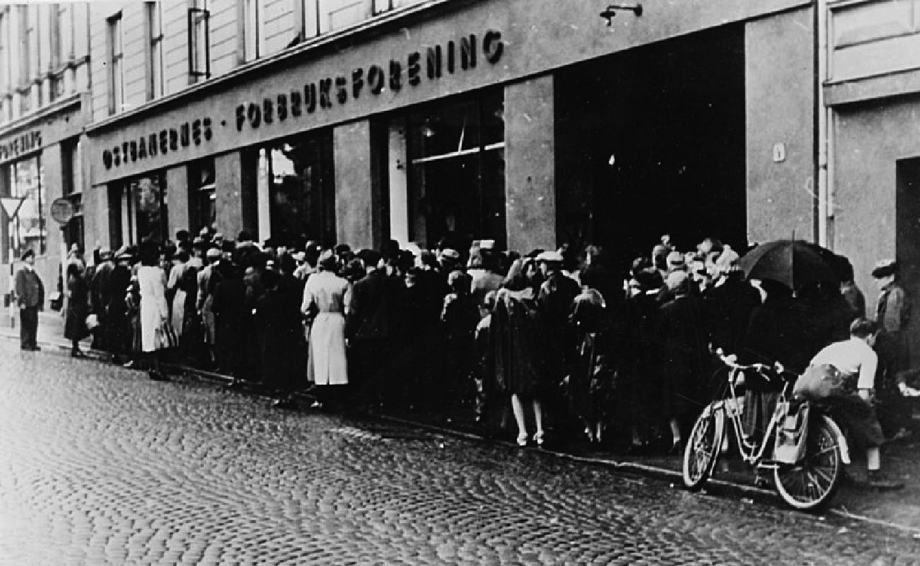 This black and white photo shows people lined up on the street in front of a building. The people are wearing coats and hats. Women wear dresses and heels. A man in front in a coat, pants, and hat observes the line. Everyone is standing very close together and the line extends the length of the building. The street in front of the line is cobblestone. There is an open umbrella at the end of the line and a boy is sitting on the wheel of his bicycle at the end of the line talking with others.