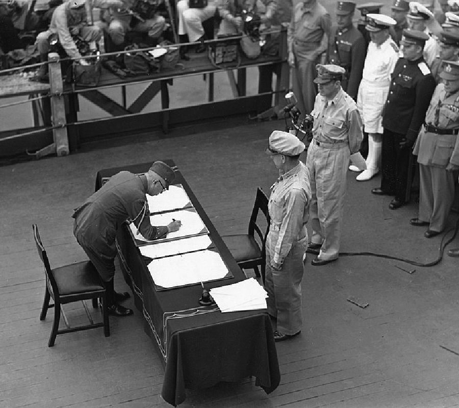 This is a black and white photograph of a man who is wearing a military uniform with tassels on his right sleeve, a hat and glasses. He stands over a desk in the middle of the picture and bends down to sign a large white paper on a rectangular table with a tablecloth that drapes over the sides, covering the table. There are four of these large papers across the table with a smaller stack of white papers on the right corner of the table. He has a chair behind himA chair is located on the opposite side of the table from him. Next to that chair stands a man who is wearing a military uniform with a hat. He watches the signing. Behind him there is a man in a military uniform standing in front of microphones. There is a group of military men in various uniforms standing in the right of the picture, watching the signing. In the top left of the picture there are more people standing and sitting looking over a railing, some taking pictures.