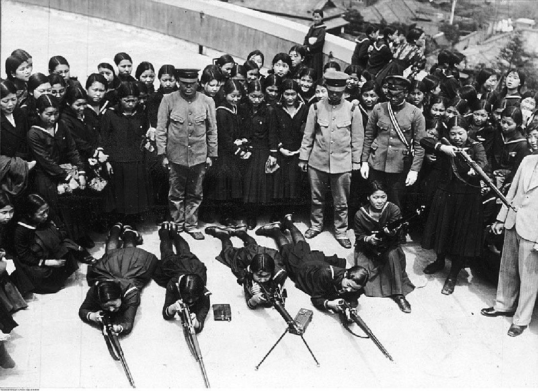 In this black and white picture, four women are laying on the floor, propped up on their elbows, looking through rifles. They are wearing dresses, stockings, and shoes. One woman is to the right of them down on one knee in a dress, holding a rifle aimed up. There is a woman in a dress to her right standing up and aiming her rifle straight out. Surrounding the women are three Japanese military personnel in uniforms, looking down at the women. Behind them all in a semi-circle are standing many other women in dresses observing. In the background on the right there is a short wall and five of the women are looking over the wall into the trees and land behind the wall. Another woman is leaning against the wall looking at the group of women standing watching those with the rifles.