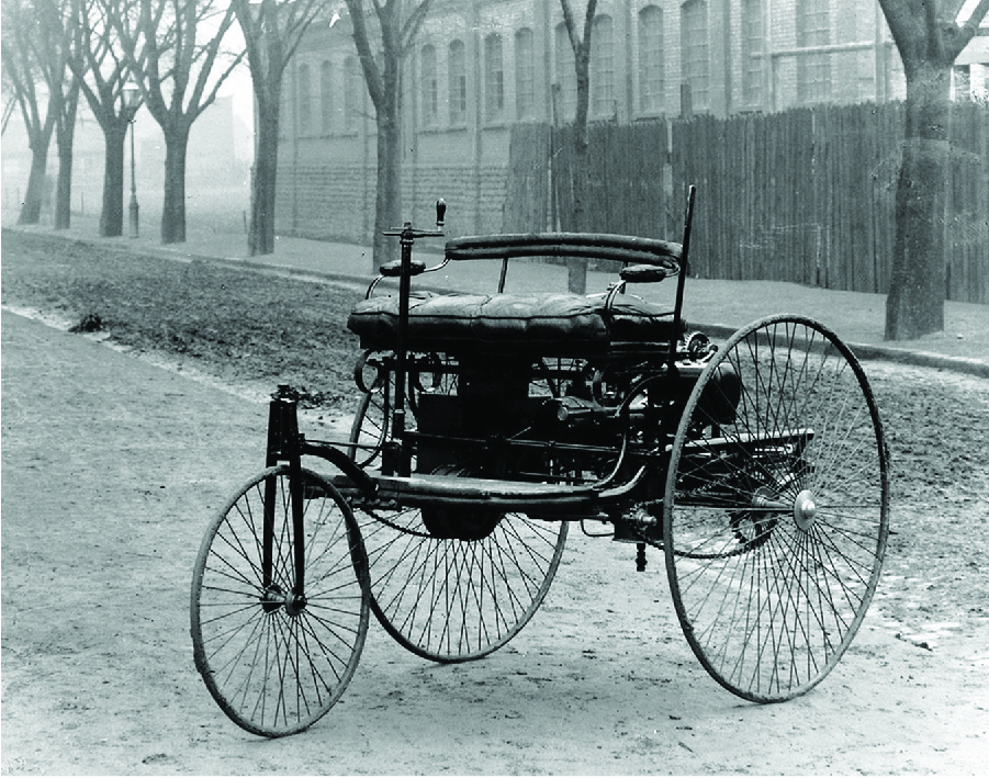 An old photograph shows an automobile. It has two tall thin wheels in the back and one smaller thin wheel at the front with many very thin spokes on all three. A floor sits high off the ground and a two-person cushioned seat with a back rail and side arms sits on that floor. A crank is on the car’s right side in front of the seat. There is gadgetry between the seat and the floor. The car is on a road with a building, a wooden fence, and trees shown in the background.