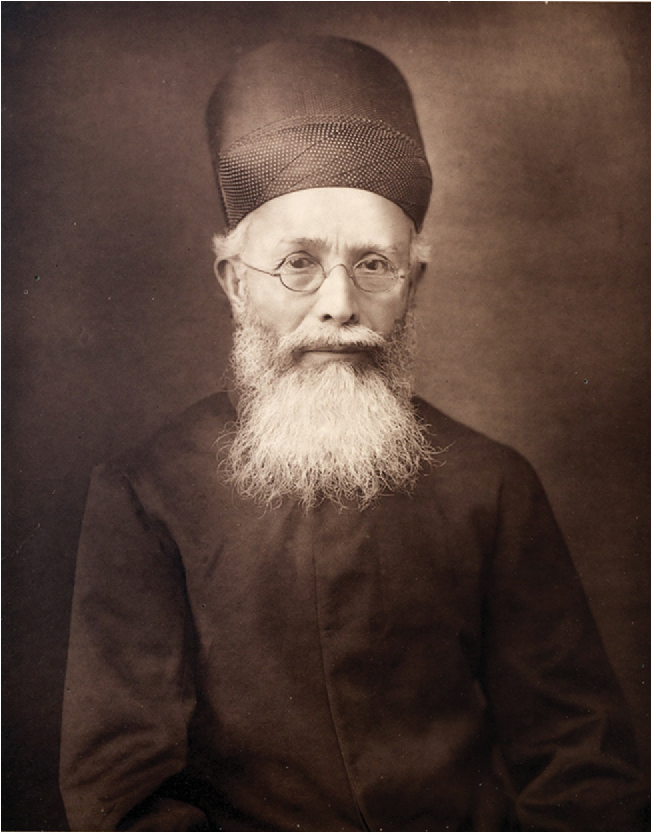 A portrait of a man with a long white beard and small round glasses. He wears a long black shirt and has a dark tall turban on his head.