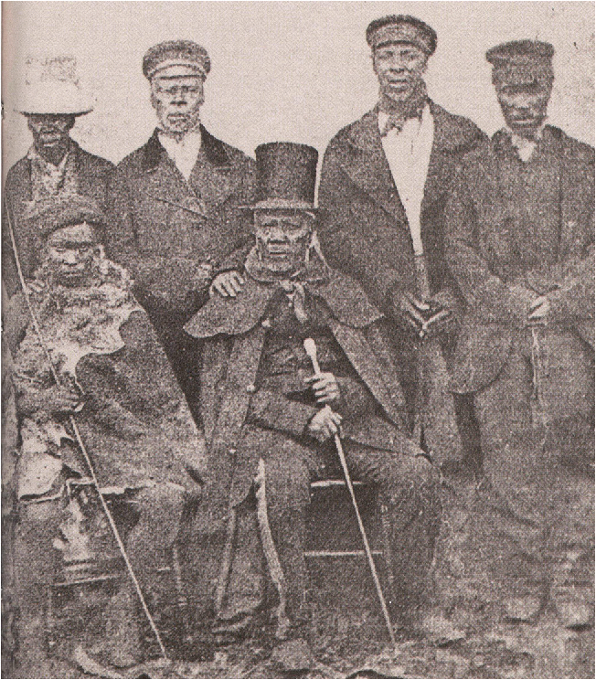A photo shows two men sitting in chairs. The one on the left wears a flowy cape, a simple hat on his head, and holds a long stick. The man to the right wears a coat, pants, top hat, a long cape coat, and holds a long cane. Behind them stand four men in long coats, and pants, the one on the far left wearing a white large hat while the other three wear smaller dark caps.