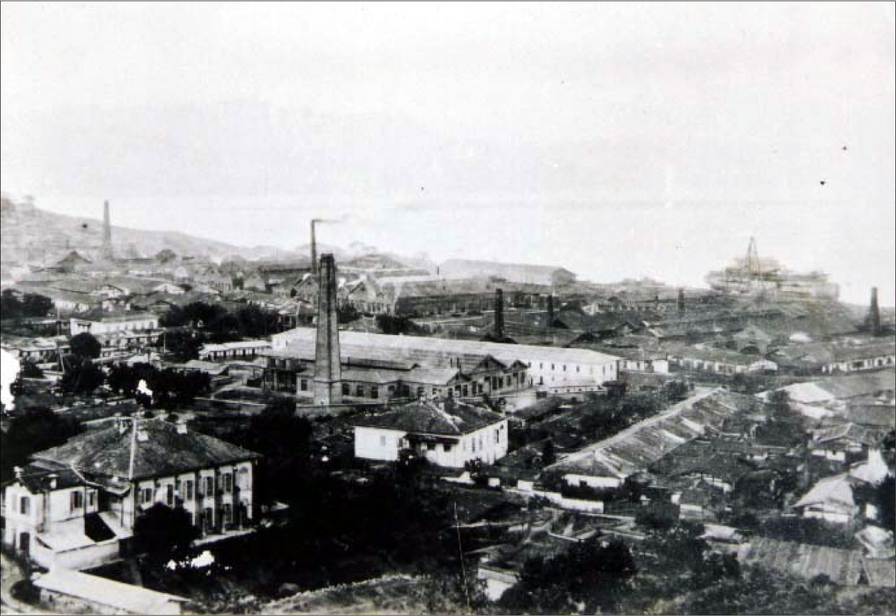 A photograph shows a tall tower in the middle surrounded by short buildings of various sizes and positioned in various direction throughout the picture. Smoke stacks are dispersed throughout the photo and a large ship is shown in the top right of the photo. Hills are seen in the back left.