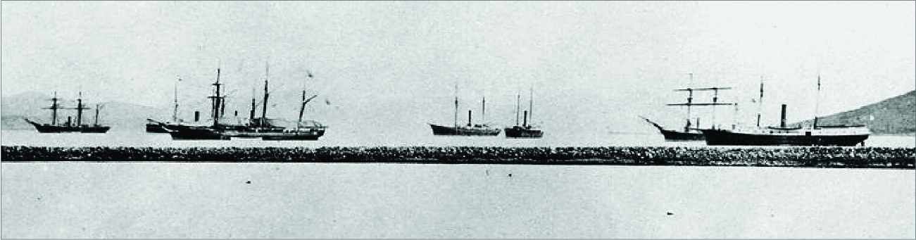 A photograph shows seven ships with tall masts in water. They have black hulls and no sails up. A stone breakwater is shown in the foreground and the background shows small hills.