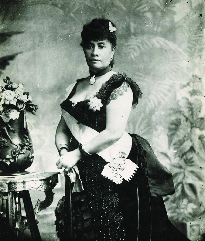 A woman is shown in a black and white photograph. She is wearing a low cut beaded dark dress with lacy cap sleeves. A brooch hangs from a beaded ribbon around her neck, her hair is up, adorned with a butterfly pin and she wears earrings, rings, and bracelets. A white sash runs from her right shoulder and ends at her left waist with a circular ornament. She holds white gloves in her hands and leans on a cane. In the left of the picture is a table with a round, carved vase filled with flowers. The wall behind her is decorated with leaves and plants.