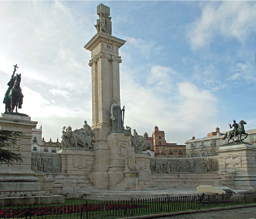 A tall tower, inscribed with “1812,” stands at the center of the monument. Several stone statues of people on foot and on horseback appear nearby.
