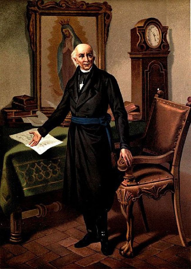 Miguel Hidalgo y Costilla points at a document that is on his desk. A painting of the Virgin of Guadalupe appears above his desk.