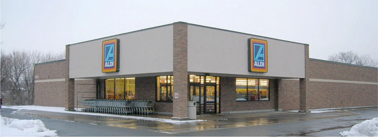 A picture shows the corner of a large, bricked building. The two sides shown are brown bricked with a beige stripe of bricks toward the top. Three windows are shown toward the front with doors in the middle. A beige flat surface shows at the two sides of the corner with an orange rectangle, blue “A”, and the word “Aldi” in white on both sides. Along the left of the front there are shopping carts in two rows along the front wall and on the right side of the middle post there is a beige garbage can. Through the windows you can see lights and some store shelves. The ground outside is wet and snow is seen in the two bottom corners. There are some trees and gray skies in the background.