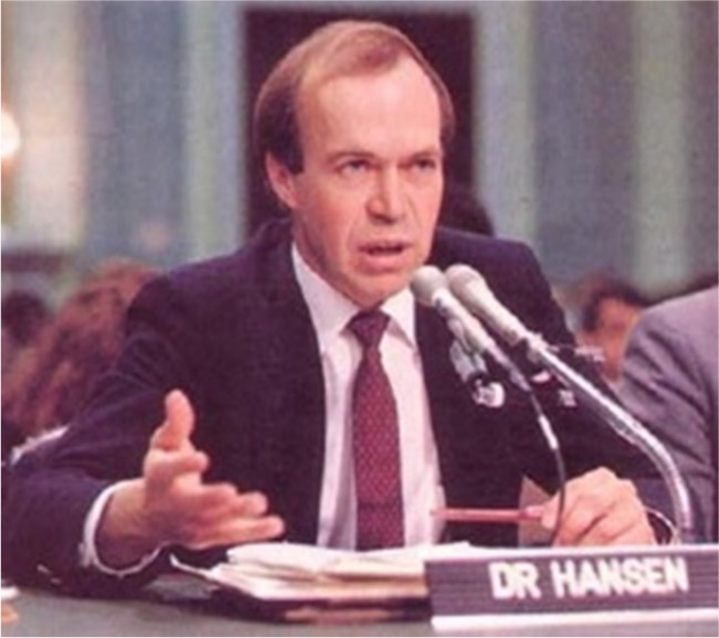 A picture is shown of a man sitting at a table in front of two silver microphones with his mouth partially open. There is a stack of papers in front of him. He is wearing a blue suit jacket, white shirt, and red tie. He has brown hair. His right hand is open and he is holding it above the table and there is a nameplate in front of the microphones with “Dr. Hansen” written on it. Behind him there is a blue wall and a head of a man and a woman can be seen as well as another man’s shoulders.