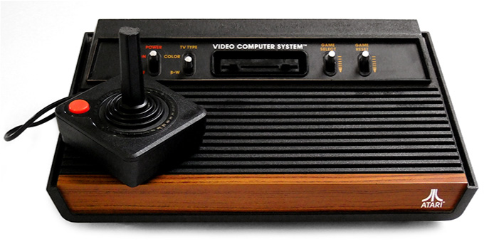 A picture is shown of a black rectangular item with a brown wooden front. There are long grooves along the front on top and a raised area along the back with switches, two on each side and a slot in the middle. The switches on the left are labeled “Power” in red and “TV Type” in yellow. “Color” and “B-W” are indicated at the top and bottom of the “TV Type” switch. The switches on the right are labelled “Game Select” and “Game Reset” in yellow with yellow arrows pointing down on the right sides of the switches. The words “Video Computer SystemTM” are written in white above the middle slot. A black square with a tall black joystick handle sits on the top left corner. It has a red button in the left corner and is connected to the console with a wire. The word “Atari®” is written in white on the bottom front of the brown panel with a symbol of one line and a curved line on either side printed above it.