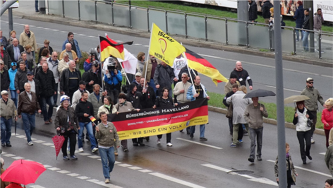 A picture of a large group of men and women walking on a wet street is shown. They wear pants, jackets, shoes, and hats. Four have umbrellas. Four people at the front of the group hold a large banner that is black, red, and yellow striped. The words “Burgerbundnis Havelland” are written in white in the top black strip, “Gewaltfrei – Unabhangig – Parteilos” are written in the middle red stripe in white and in black on the bottom yellow stripe are the words “-Wir Sind Ein Volk –“ in the middle with “Burgerbundnis Havelland” in smaller black letters on both sides. A white “F” in a blue box is also seen at the right. Behind the banner four people carry large flags. Two flags are black, red, and yellow striped while one is white with some red and one is yellow with the words “Islam” and “Neinn” seen, with the rest obscured. In front of the banner twelve people are standing and walking about, some with umbrellas, some hugging, some talking. In the background of the picture, there is an opaque gate on the curb with grass behind and six people standing, watching, and walking. Parts of a white building can be seen behind the people.