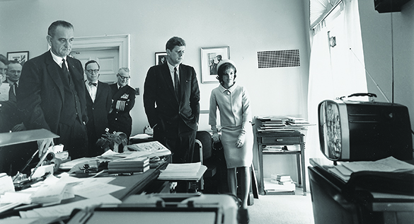 Una fotografía muestra a John F. Kennedy, Jacqueline Kennedy, Lyndon Johnson y varios otros de pie en una oficina de la Casa Blanca, viendo una pequeña televisión.
