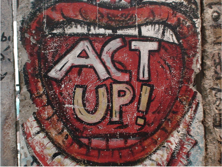 A picture of three sections of a concrete wall is shown. A mural of an open mouth is drawn on the larger middle section. It has red lips, a red painted inside, and teeth, white on top and red at the bottom. The words “Act Up!” are painted inside the mouth, “act” in white and “up!” in tan. The side sections of the well have dark scribblings on them.