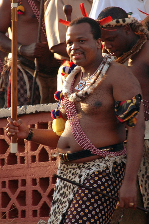 A picture of a dark-skinned man with dark hair and a horse shoe-shaped, black moustache is shown. He wears no shirt, has various leis and necklaces around his neck, and tassels in bright yellow, blue, and orange hang on his right arm. Eight rows of pink beads hang across his chest from his right shoulder to his waist at the left. The top of his chest has brown skin tags all over and his belly protrudes over a black belt with a gold buckle. Black and white animal print fur hangs below his waist over a black cloth with orange and white circles. On the underside of his right wrist a black watch band can be seen and he is holding a tall brown stick with a white bottom and white ring around the top. The top of his left arm is adorned with a black, yellow and orange cloth and he wears a gold plain ring on the third finger of his left hand which holds a thin dark stick. A dark-skinned man stands behind him with an open mouth and his head hanging down. He has dark hair, wears a black and white animal fur around his head with orange feathers coming off the front and several necklaces and adornments around his neck. He holds a tall stick with a rounded top. Behind both men a red brick wall stands with white trim at the top and three other people are shown standing behind it, two in light-colored short sleeved shirts and one with a bare chest, beads, and a patterned cloth below the waist holding a stick in his hand.