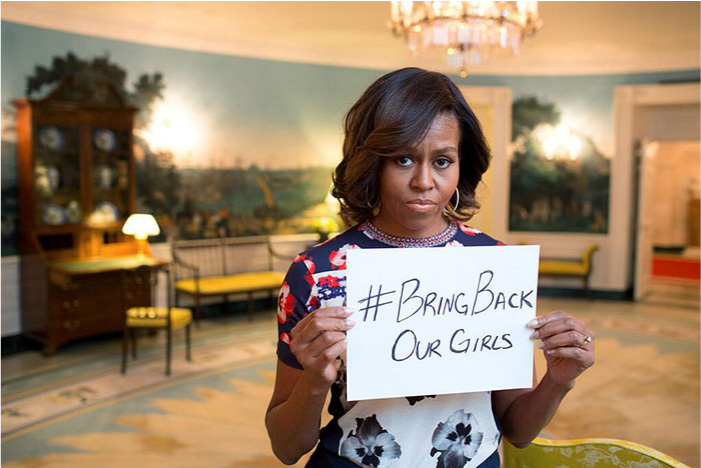A picture shows an African American woman standing holding a small white paper with “#Bring Back Our Girls” written on it in black ink. She has short wavy hair, large hoop earrings, a blue, red, and white flowered dress, and a diamond ring on her left hand. In the background a blurry room is shown with a tall desk, chair, benches, a lamp, and walls decorated with paintings of scenery. A large, glass chandelier hangs above her head in the background.