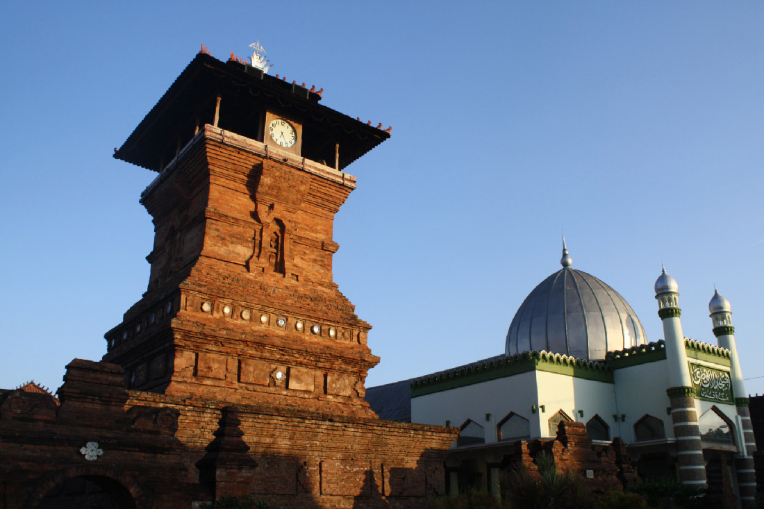 An image of two buildings is shown. The building on the left is a square, tall tower, made from red brick, with a clock near the top. There are minimal decorative carvings on the front and circular and square decorations along the bottom. The building on the right has a large, silver dome with a short spire and two smaller dome striped and decorated towers at the front. Green scalloped trim runs along the edges of the roof and pointed windows run along the bottom of the building.