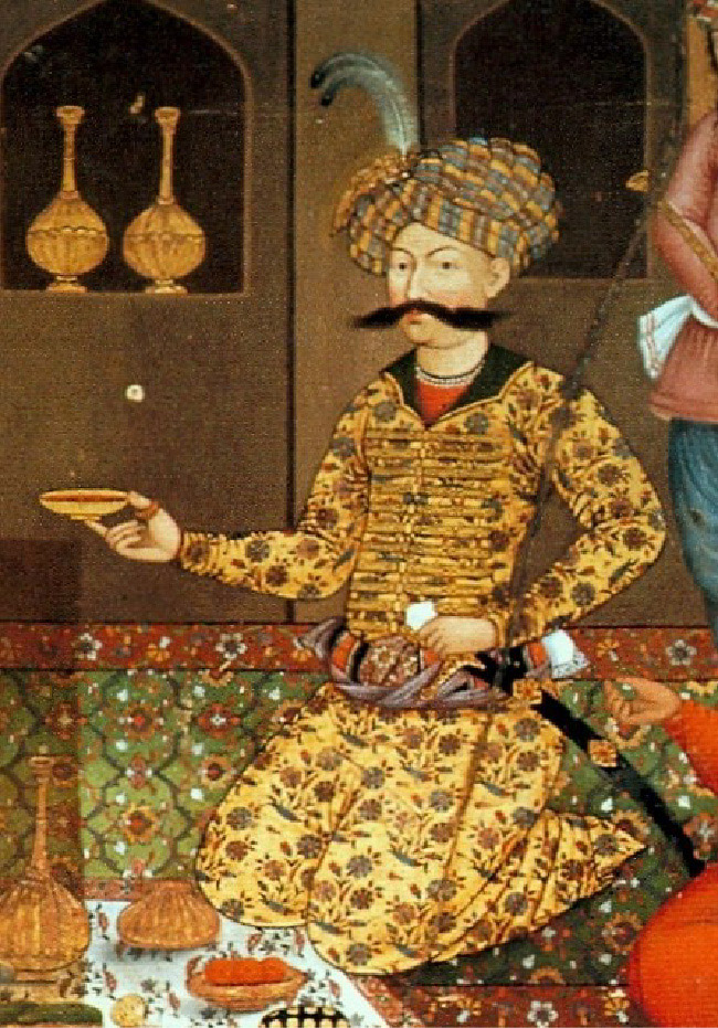 A painting of a kneeling man on a lavishly decorated rug is shown. He wears a striped woven turban with a white feather in the front. His long robe has flowers all over and a red shirt shows by his collar. He has a long moustache and an ornate belt around his waist. His left hand rests on the handle of a sword and his right hand holds a dish. A cloth in the bottom left of the painting holds vases and bowls while the arms and legs of two people can be seen in the right edges of the painting. The wall behind him displays two gold vases on a ledge.