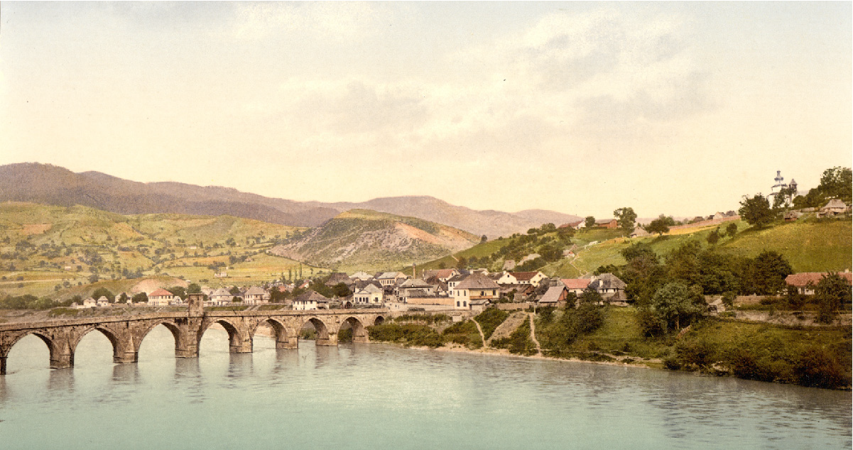An image of a stone arched bridge over water is shown. Buildings are on the hills surrounding the water. Hills with trees and farmland are visible in the background.