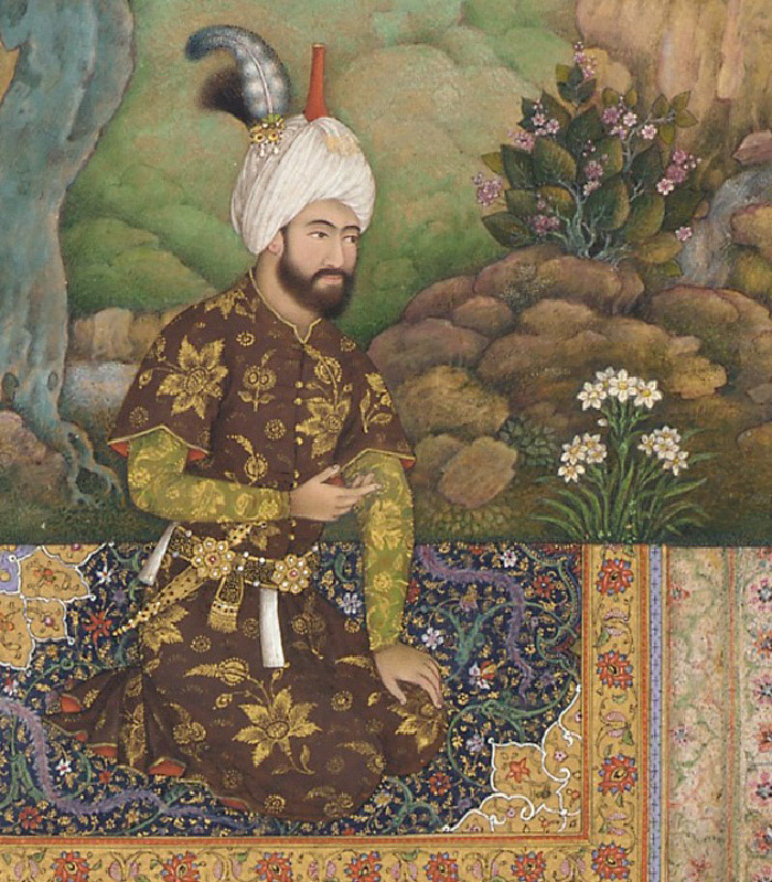 A painting of a bearded man kneeling on a richly decorated rug in a garden is shown. He is wearing a brown short sleeved long coat embossed with gold flowers over a green long-sleeved shirt with gold flowers. He wears a white turban on his head with a red projection at the top and a silvery feather in the back. His belt holds a short sword in a sheath and objects dangling from elaborate flowery decorations. In the background a tree, flowers, rock and grass can be seen.