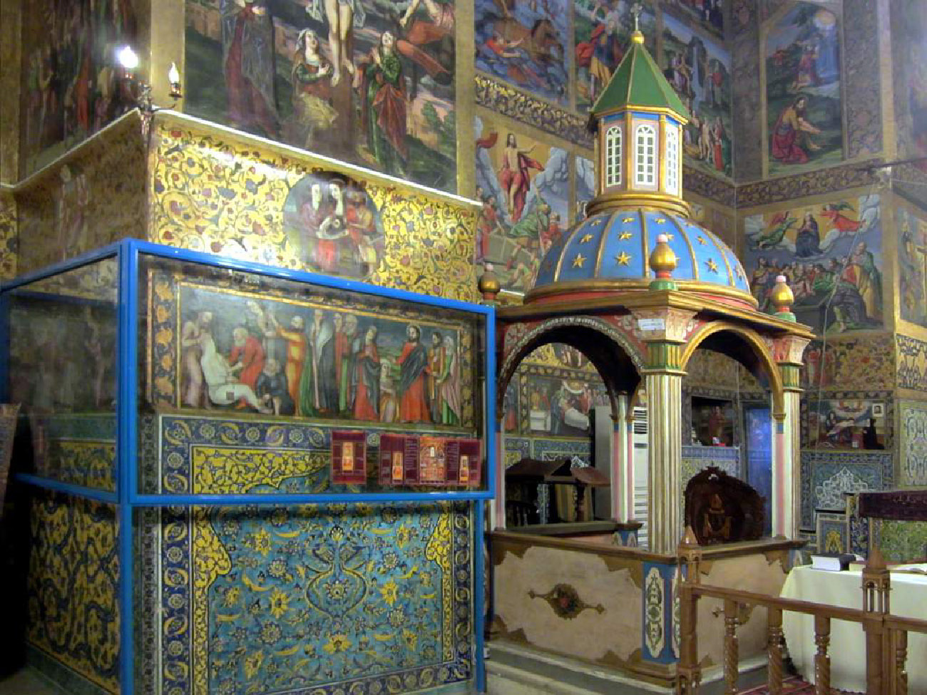 A photograph of an interior of an elaborately decorated room is shown. A small gazebo is located in the right portion of the photograph with a blue starry domed top and a steeple with white grates and a green roof. Part of an altar is shown to the left. The walls of the rest of the room are filled with paintings of people in long robes, women and children, as well as other religious scenes. Around the paintings the walls are decorated with flowery tiles.