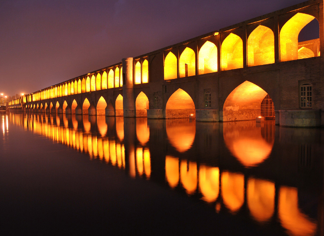 A photograph of a bridge with arched sections at the bottom and top is shown at night. Lights reflect from inside the top portion and the entire bridge reflects in the water below.