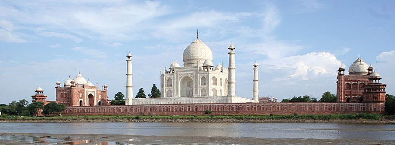 The Taj Mahal is in the center of the photograph. A mosque sits on either side of the Taj Mahal. The Yamuna River is visible in the foreground.
