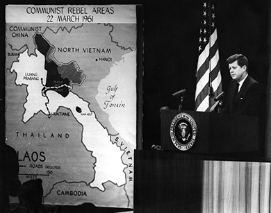 Una fotografía muestra al presidente Kennedy parado en un podio pronunciando un discurso. A su lado cuelga un gran mapa del sudeste asiático, etiquetado como “Áreas rebeldes comunistas/22 de marzo de 1961”.
