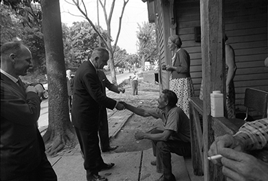 Una fotografía muestra al presidente Johnson parado en una calle afuera de una casa, varios de cuyos habitantes se sientan y se paran en el porche. Le da la mano a un hombre sentado mientras otros dos funcionarios miran.
