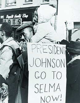 一张照片显示一群非裔美国人在阿拉巴马州塞尔玛的街道上游行。 在前景中，一个肩上挂着小孩的男人举着一个标语，上面写着 “约翰逊总统/现在去塞尔玛吧！”