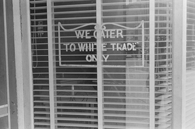 Une photographie montre une vitrine portant une pancarte sur laquelle on peut lire « Nous nous adressons uniquement au commerce blanc ».