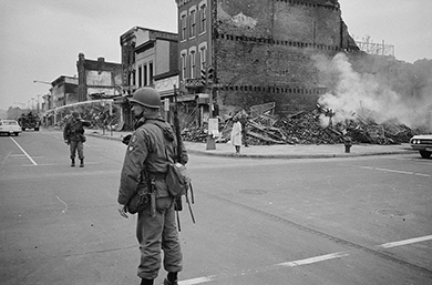 一张照片显示了一条街道，空无一人，但有一名行人和几名身穿防暴装备的男子。 在拐角处可以看到建筑物的废墟。