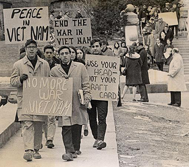 Uma fotografia mostra estudantes protestando no campus da Universidade de Madison-Wisconsin. Eles seguram cartazes com os dizeres “Chega de guerra no Vietnã”; “Paz no Vietnã”; “Acabe com a guerra no Vietnã”; e “Use sua cabeça, não seu cartão de seleção”.