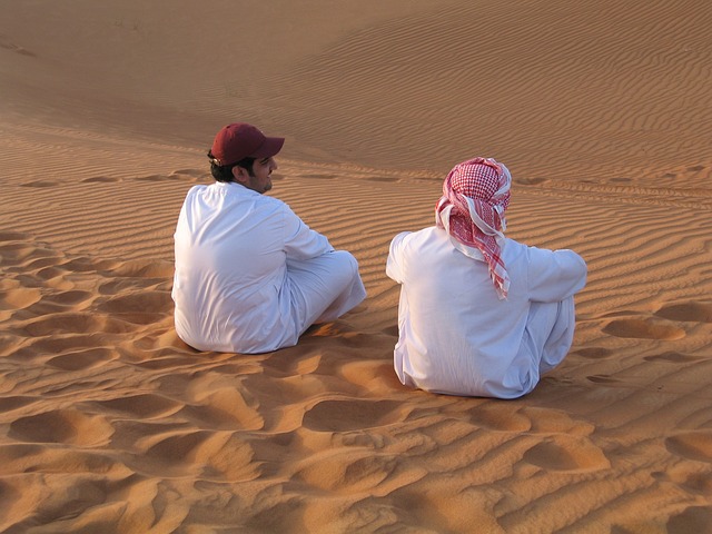 Two male friends are chatting while sitting on the dessert sand. 