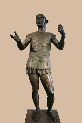 This is a photo of the statue the Mars of Todi. This bronze statue depicts a male Etruscan warrior wearing intricate body armor. His pose places most of his weight on one foot so that his shoulders and arms twist off-axis from his hips. His left hand is closed, as if gripping an object. His right hand is outstretched as if offering an object.