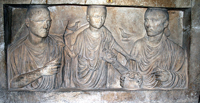 This is a photo of a relief of a Roman family. The father (left), the child (center), and the mother (right). The family wears Roman togas. There is a bird perched on the father's arm, which rests on the child. A second bird is in the sky above the child's right shoulder.
