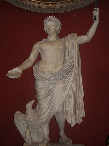 This photo shows a statue of Claudius as Jupiter. Claudius wears a laurel crown and a toga, which reveals his idealized chest and torso. An eagle-like bird rests by his foot.