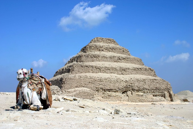 Photograph depicts tiered mud brick pyramid against a blue sky. In the bottom left corner of the shot, there is a camel lying down.