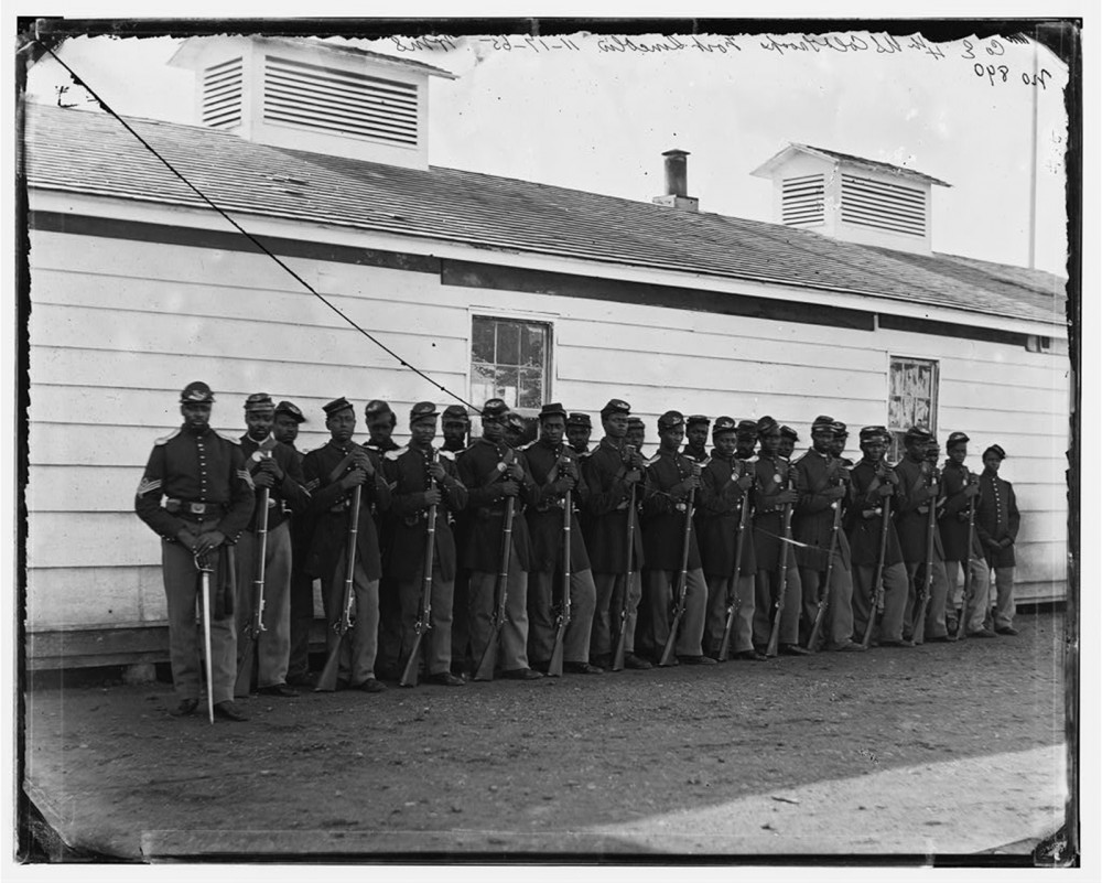 This photograph shows a group of Black soldiers, standing at ease in uniform with their rifles.