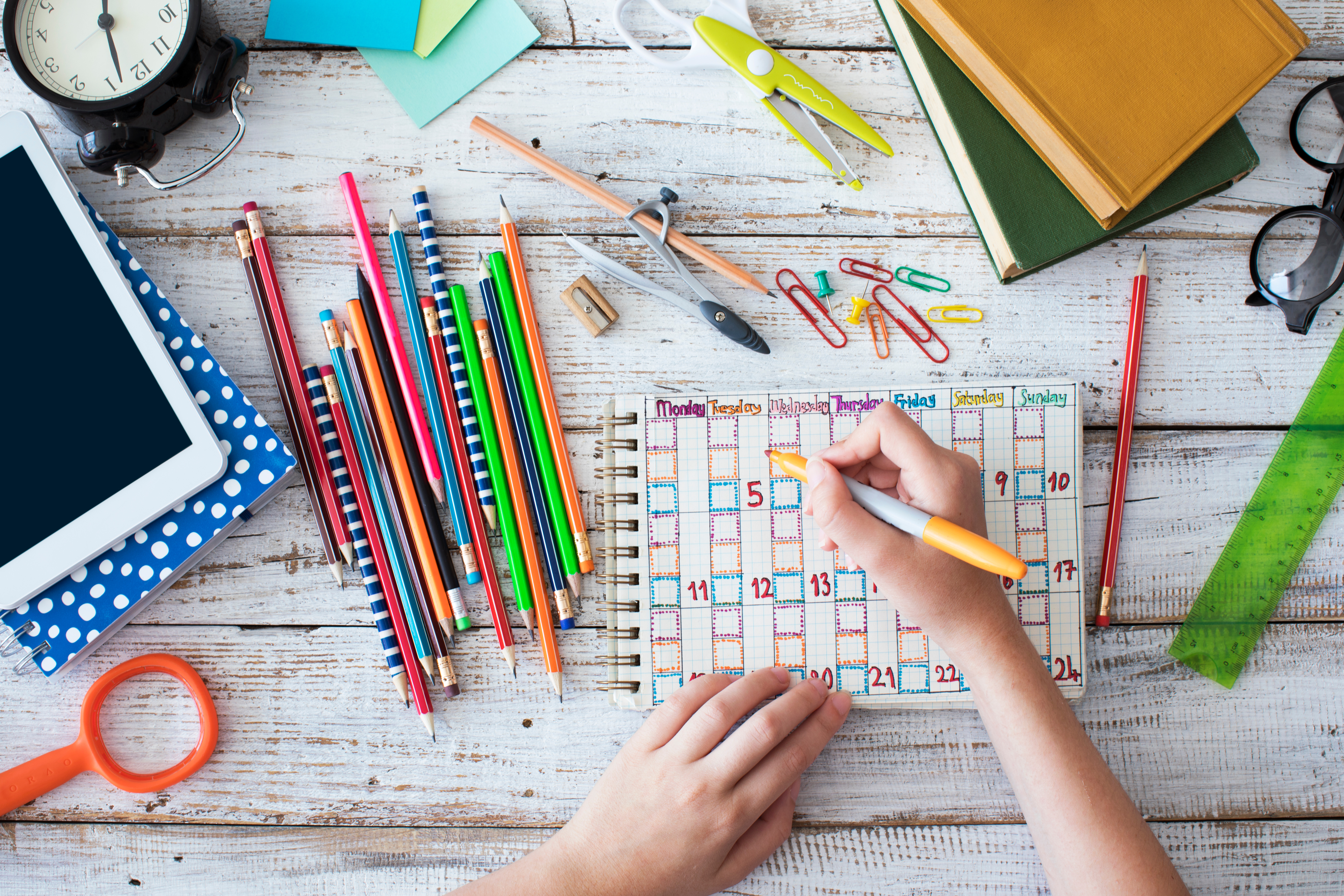 A person writes in a colorful planner on a wooden desk cluttered with colored pencils, paper clips, books, a tablet, scissors, glasses, and a ruler.