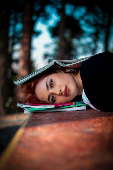 A student rests her head on a stack of colorful textbooks with an open book on top, outdoors with a blurred tree background.