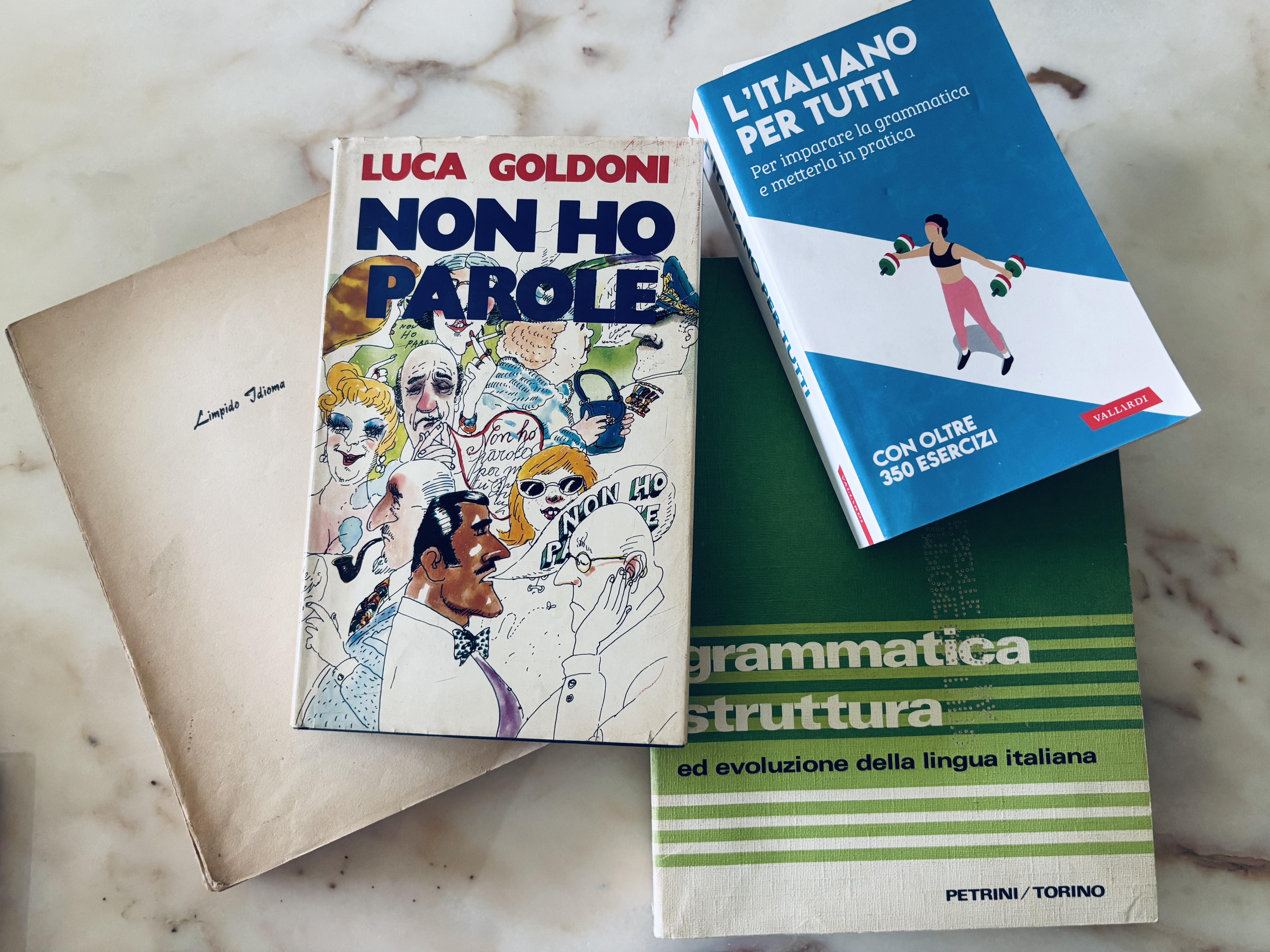 Four books on a marble surface: a beige book titled "Non ho Parole" by Luca Goldoni, a green book on Italian grammar, and a blue workbook titled "L'Italiano per Tutti."