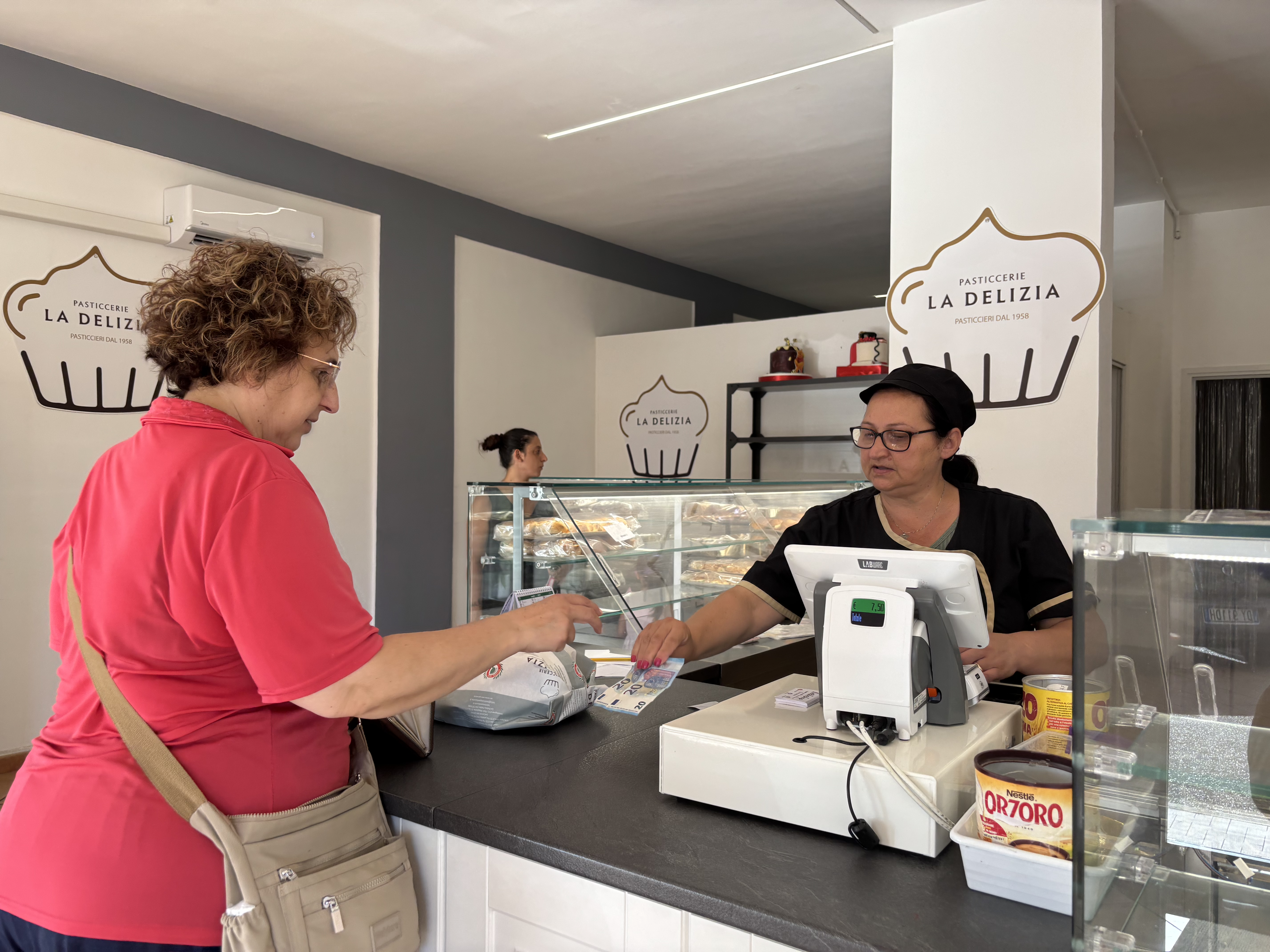 A woman is paying at the cashier in an Italian bakery. Behind the counter, the cashier is ready to accept her payment.
