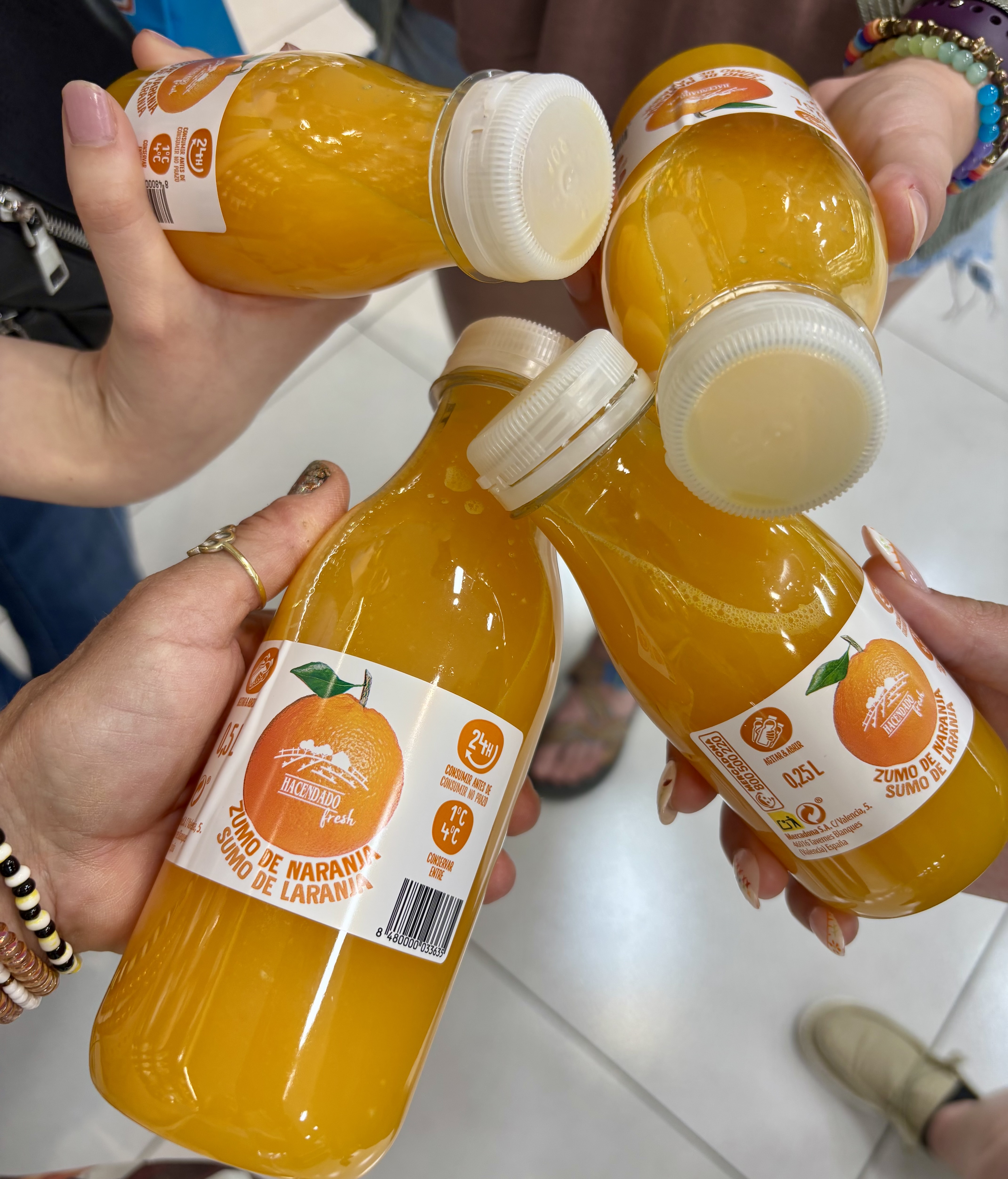 Hay una foto de cuatro manos con cuatro botellas de jugo fresco de naranja.