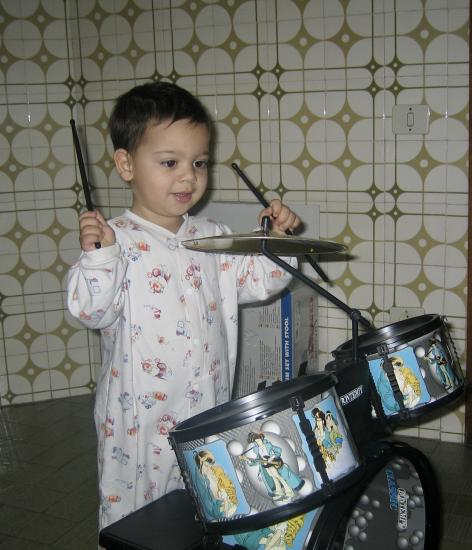 A young child, about three years old, happily playing a toy drum with two drumsticks.